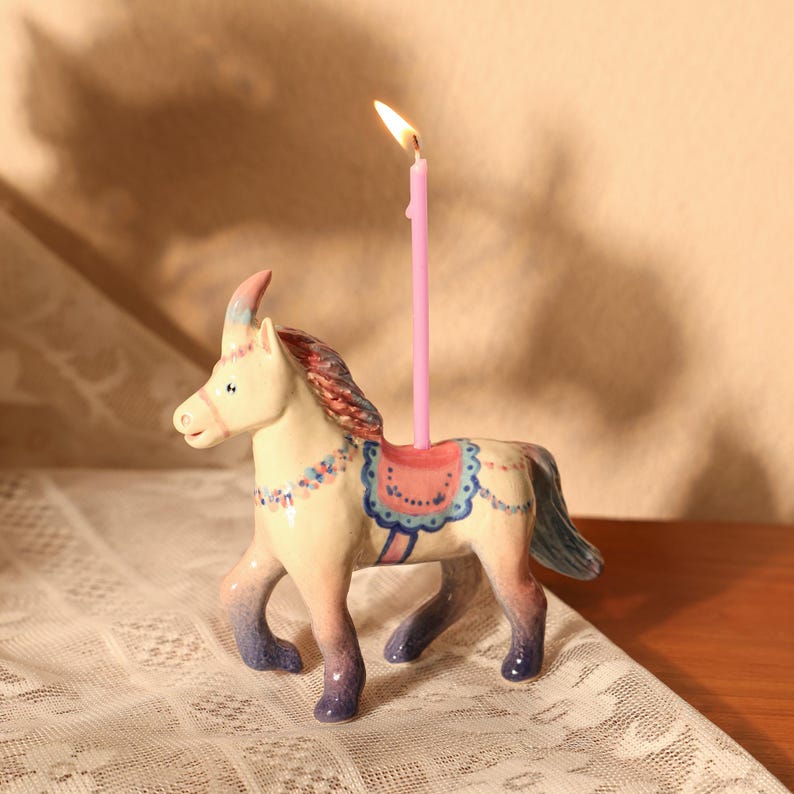 Op de afbeelding: Een keramieken paardenbeeldje met een roze kaars. Het paard is geschilderd in pastelkleuren met blauwe, roze en cr&egrave;mekleurige accenten. De kaars is aangestoken en het beeldje heeft een zadelontwerp.