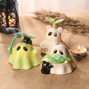 May include: Three ceramic ghost figurines with leaf accents. One is light green with blue dots, another is white with a leaf, and the third is white with a sprout. A lit tealight candle and a decorative lantern are also present.