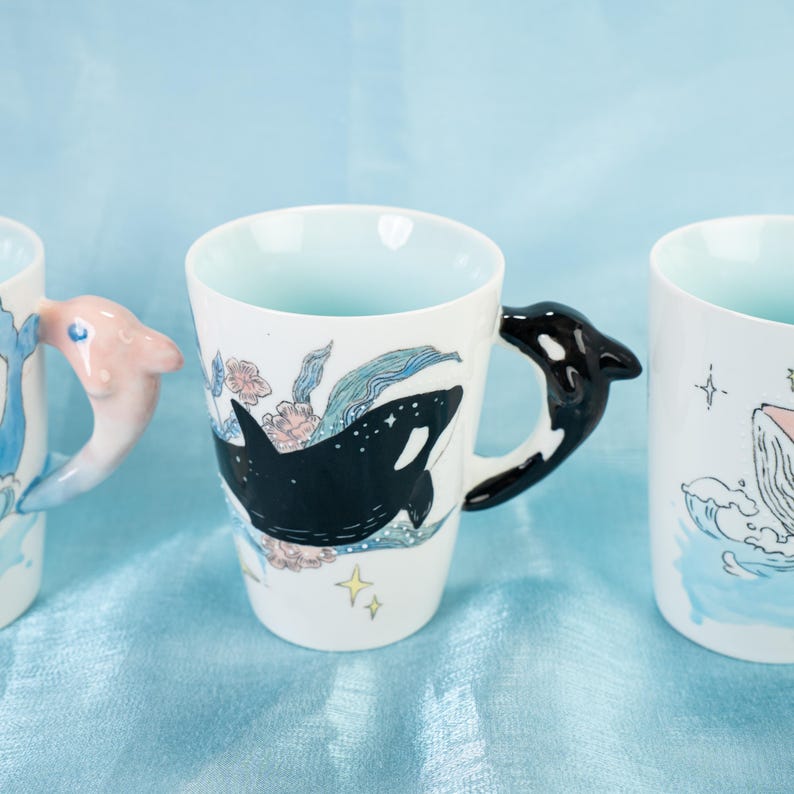 Puede incluir: Tres tazas de cer&aacute;mica con dise&ntilde;os de tem&aacute;tica oce&aacute;nica. La taza de la izquierda tiene un asa de delf&iacute;n rosa, la taza del medio tiene un asa de orca negra y la taza de la derecha tiene un asa de ballena azul. Cada taza presenta un animal marino diferente y dise&ntilde;os florales.