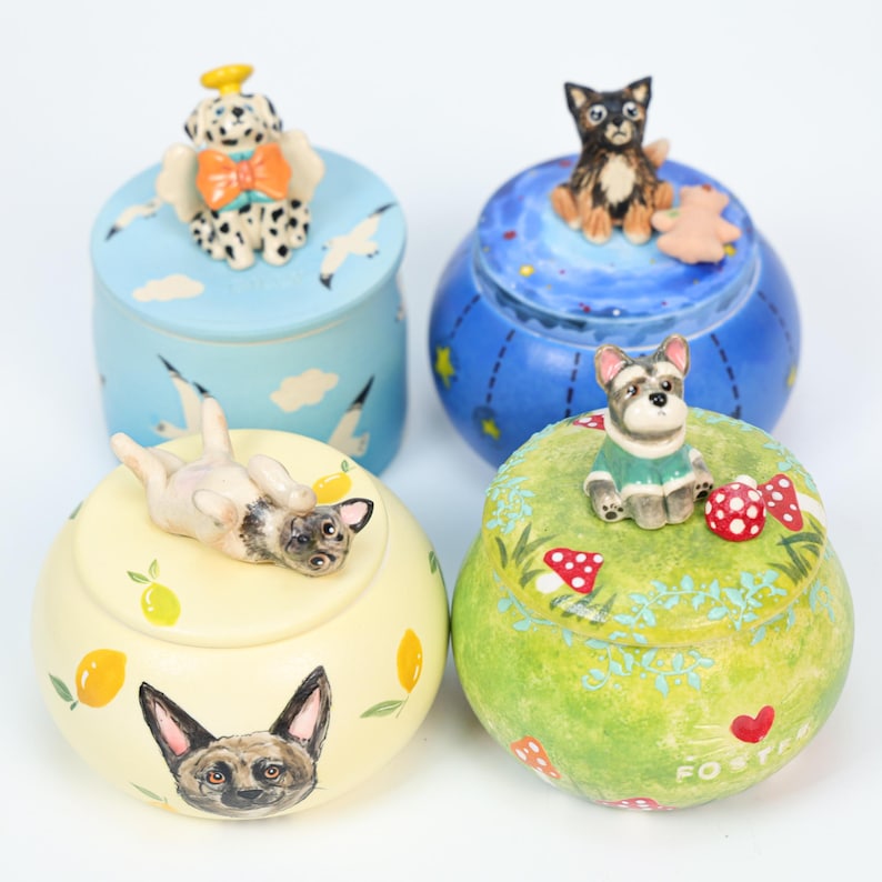 May include: Four decorative ceramic jars with dog-themed designs. One jar is light blue with a Dalmatian angel on top, another is blue with a dog and a teddy bear, a third is yellow with a pug, and the last is green with a dog.