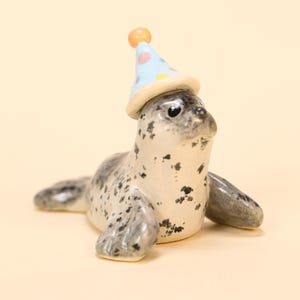 May include: A ceramic seal figurine with a light blue party hat adorned with pastel polka dots. The seal is primarily white with black spots and gray flippers, set against a cream-colored backdrop.