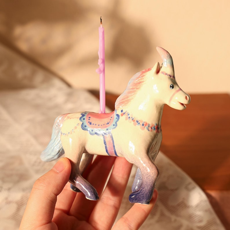 Op de afbeelding: Een keramieken eenhoornbeeldje met een roze kaars. De eenhoorn is geschilderd in pastelkleuren, met een blauwe en roze manen en staart, en een blauw zadel. De kaars is roze en is aangestoken. Het beeldje wordt in een hand gehouden.