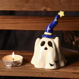 May include: A ceramic Halloween ghost candle holder with a blue wizard hat adorned with yellow stars. The ghost has cut-out eyes and mouth, with two black bat silhouettes. A lit tea light candle sits nearby on a wooden surface.