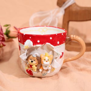 Puede incluir: Una taza de cerámica con borde rojo de lunares y asa. La taza presenta un diseño 3D de un perro y un gato que emergen de un efecto de papel rasgado. La base es blanca con detalles florales y de setas. El asa es de color canela con rayas marrones.