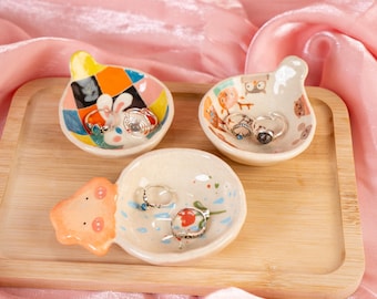 Puede incluir: Tres platos de cerámica para anillos con diseños caprichosos. Cada plato presenta un animal diferente y patrones coloridos. Los platos están dispuestos en una bandeja de madera.