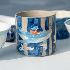 Puede incluir: Una taza de cerámica azul y blanca con un diseño caprichoso de una niña montando una ballena. La taza tiene un patrón a rayas y un borde marrón claro.