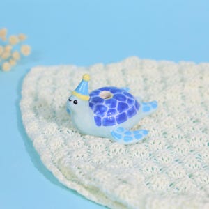 May include: A small ceramic turtle figurine with a light blue body and a dark blue patterned shell. The turtle wears a tiny yellow and blue party hat on its head and features a small circular opening on the top of its shell, finished in a glossy glaze.
