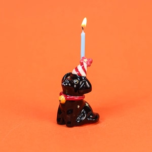May include: A black ceramic dog-shaped birthday candle holder with a lit blue candle. The dog wears a red collar with a gold bell and a party hat. The candle holder is decorated with gold stars and sits against an orange background.