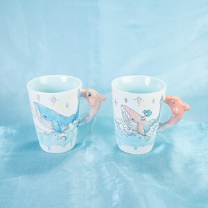 Puede incluir: Dos tazas de cer&aacute;mica blanca con un dise&ntilde;o de una ballena azul y una ni&ntilde;a montando la ballena. Las tazas tienen asas de delf&iacute;n rosa.
