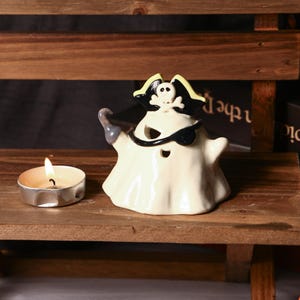Könnte beinhalten: Eine Keramik-Halloween-Dekoration in Form eines Geisterpiraten. Der weiße Geist hat eine schwarze Augenklappe, einen schwarz-gelben Piratenhut und einen grauen Haken. Eine brennende Teelichtkerze steht neben dem Geist auf einem Holzregal.