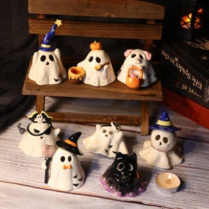 May include: A collection of ceramic Halloween figurines shaped like ghosts, each with unique accessories. Some ghosts wear hats, carry baskets, or have cat-like features. The figurines are white with black facial features and accents, set on a wooden surface.