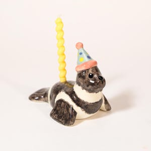 May include: A ceramic seal figurine featuring a glossy finish with black and white markings. The seal wears a small, conical party hat in light blue with colorful polka dots and a pink rim. A tall, yellow, spiral-shaped candle stands upright.