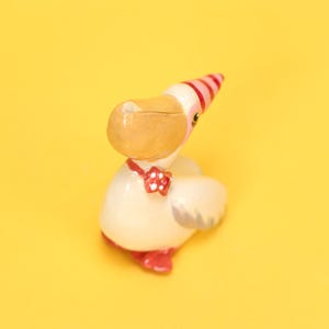 以下が含まれることがあります： 明るいベージュの体、大きなタン色のくちばし、赤と白のストライプのパーティーハットをかぶった鳥の気まぐれな置物。白い水玉模様の赤い蝶ネクタイをしており、赤い足があります。背景は無地の黄色です。