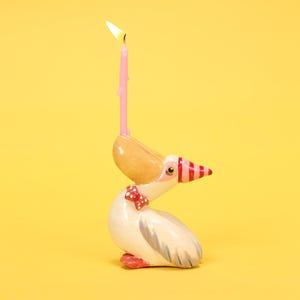 以下が含まれることがあります： 風変わりなペリカンの形をしたキャンドルホルダー。セラミック製のペリカンは、白い体に灰色の翼、赤と白のストライプのくちばし、赤い蝶ネクタイが特徴です。火のついたピンク色のろうそくがペリカンの頭に差し込まれています。背景は黄色です。