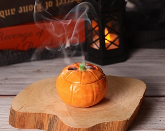 May include: An orange ceramic pumpkin-shaped incense burner with a green stem, releasing wisps of smoke. The burner is on a wooden slice, with a black lantern and books in the background. A Halloween decoration.