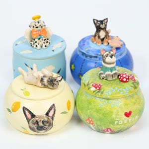 May include: Four decorative ceramic jars with dog-themed designs. One jar features a pug figurine, another has a dog angel, and two others have dog figurines on top. The jars are painted in various colors, including blue, yellow, and green.