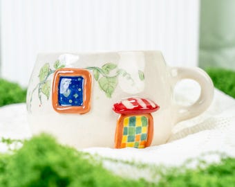 Puede incluir: Taza de cerámica blanca con un diseño de casa de ensueño. La casa tiene un techo a rayas rojas y blancas, una puerta a cuadros verdes y amarillos y una ventana azul con estrellas blancas. La taza está decorada con enredaderas verdes.