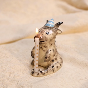 Pode incluir: Uma estatueta de dragão em cerâmica com acabamento manchado em castanho e creme, usando um pequeno chapéu de festa às riscas azuis e brancas. O dragão segura uma vela branca fina decorada com pequenas estrelas douradas, acesa com chama.