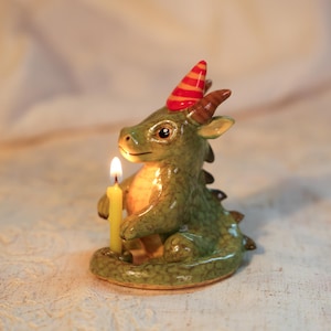 K&ouml;nnte beinhalten: Eine gr&uuml;ne Drachenfigur aus Keramik mit einer Partyh&uuml;tchen mit roten und orangefarbenen Streifen. Der Drache h&auml;lt eine brennende gelbe Kerze. Die Figur hat braune H&ouml;rner und ein detailliertes, freundliches Gesicht. Der Hintergrund ist sanft beleuchtet.