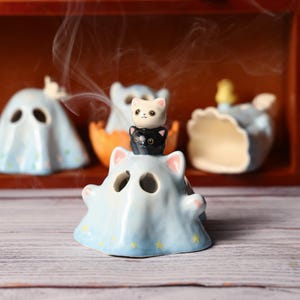May include: A collection of ceramic cat-themed incense holders. The main holder is light blue with a cat shape and star accents. A small cat figurine sits atop it. Smoke rises from the holders, suggesting use.
