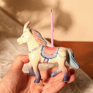 Op de afbeelding: Een keramieken paardenbeeldje met een roze kaars. Het paard is beschilderd in pastelkleuren, met blauwe en roze accenten op de manen, het zadel en de benen. De kaars is in de rug van het paard geplaatst, wat een decoratief object cre&euml;ert.