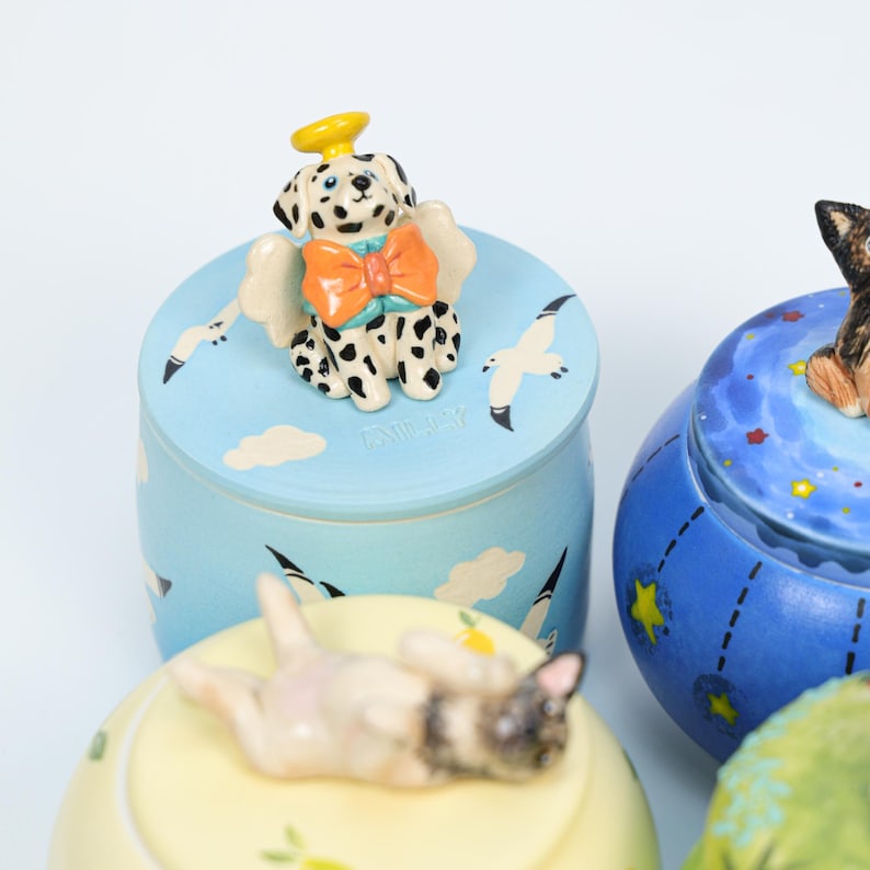May include: A collection of decorative ceramic jars with lids, each featuring a different animal figurine. One jar has a Dalmatian with angel wings and a halo. The jars are painted in various colors, including blue, yellow, and green.