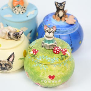 May include: A collection of decorative ceramic jars with dog-themed lids. The jars are painted in various colors, including blue, yellow, and green, and feature hand-painted dog figurines and whimsical designs. The word "FOSTER" is visible on one jar.
