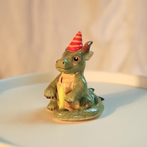 K&ouml;nnte beinhalten: Keramik-Drachenfigur mit gr&uuml;nem K&ouml;rper, gelbem Bauch und braunen H&ouml;rnern. Der Drache tr&auml;gt einen rot-orange gestreiften Partyhut und h&auml;lt eine gelbe Kerze. Die Figur steht auf einer wei&szlig;en Oberfl&auml;che.