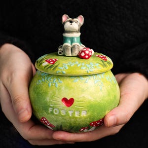 May include: A green ceramic jar with a lid featuring a dog figurine and red mushroom accents. The jar has the word "FOSTER" and a red heart design. The jar is held in a person's hands.