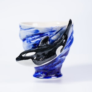 Könnte beinhalten: Eine blaue und weiße Keramiktasse mit einem Design aus einem schwarzen und weißen Orca-Wal. Die Tasse ist wellenförmig und der Orca schwimmt durch die Welle.