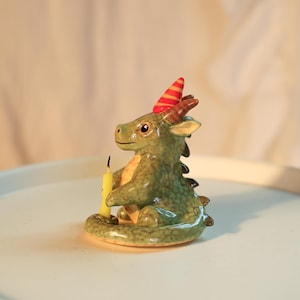 K&ouml;nnte beinhalten: Eine gr&uuml;ne und gelbe Keramikdrachenfigur mit roter und gelber Partyh&uuml;tchen. Der Drache h&auml;lt eine gelbe Kerze. Die Figur steht auf einer wei&szlig;en Oberfl&auml;che.