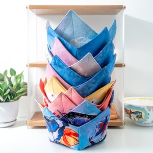 Può includere: Una pila di sei scaldavivande in tessuto colorati in una varietà di motivi, tra cui disegni floreali, geometrici e astratti. Gli scaldavivande sono disposti a forma di piramide su uno scaffale di legno.