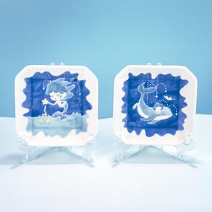 Op de afbeelding: Twee witte vierkante keramische borden met blauwe en witte illustraties van een zeemeermin en een walvis. De borden staan op heldere plastic standaards.
