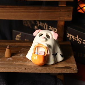 Puede incluir: Decoración de Halloween de cerámica con forma de perro fantasma que sostiene una cesta naranja llena de caramelos. El perro tiene orejas y nariz rosas. Murciélagos negros decoran la parte inferior del fantasma. Un cono de incienso está a la izquierda.