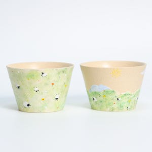 May include: Two decorative ceramic bowls. One bowl features a green background with sheep and flowers. The other bowl has a beige background with a sun, clouds, and sheep. The bowls have a light beige interior.