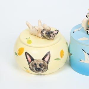 May include: A yellow ceramic jar with a lid featuring a small dog figurine lying on its back. The jar is decorated with a dog's face and lemon illustrations. A second blue jar with a bird design is partially visible.