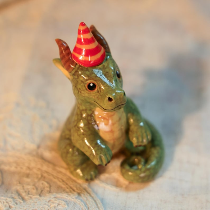 K&ouml;nnte beinhalten: Keramikfigur eines gr&uuml;nen Drachen mit einer Partyh&uuml;tchen mit orangefarbenen und gelben Streifen. Der Drache hat braune H&ouml;rner und einen wei&szlig;en Streifen auf dem Kopf. Die Figur steht auf einer strukturierten Oberfl&auml;che.
