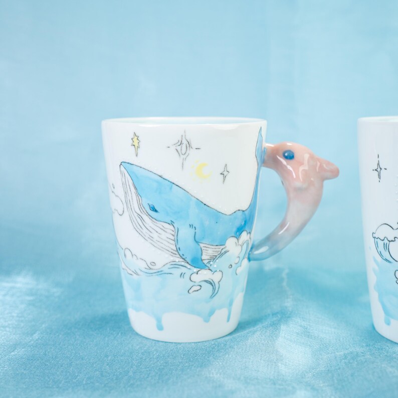 Puede incluir: Una taza de cer&aacute;mica blanca con una ilustraci&oacute;n de una ballena azul y un asa de delf&iacute;n rosa. La taza tiene un dise&ntilde;o de acuarela con olas azules y blancas y estrellas.
