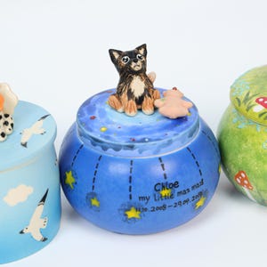 May include: A collection of decorative ceramic jars. The central jar is blue with a painted night sky design, a small dog figurine, and the text "Chloe my Little maz maz 11.10.2008-29.04.2019". Other jars are light blue and green.