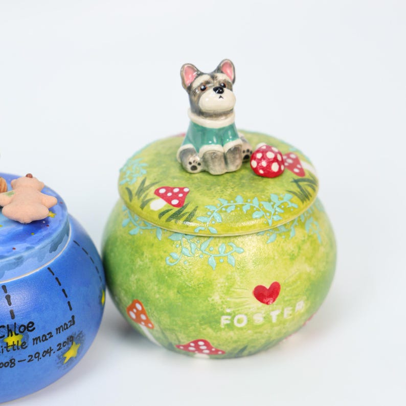 May include: A ceramic jar with a green lid featuring a Schnauzer figurine, red mushrooms, and the name "FOSTER". The jar has a green and yellow base. The jar is a decorative item.