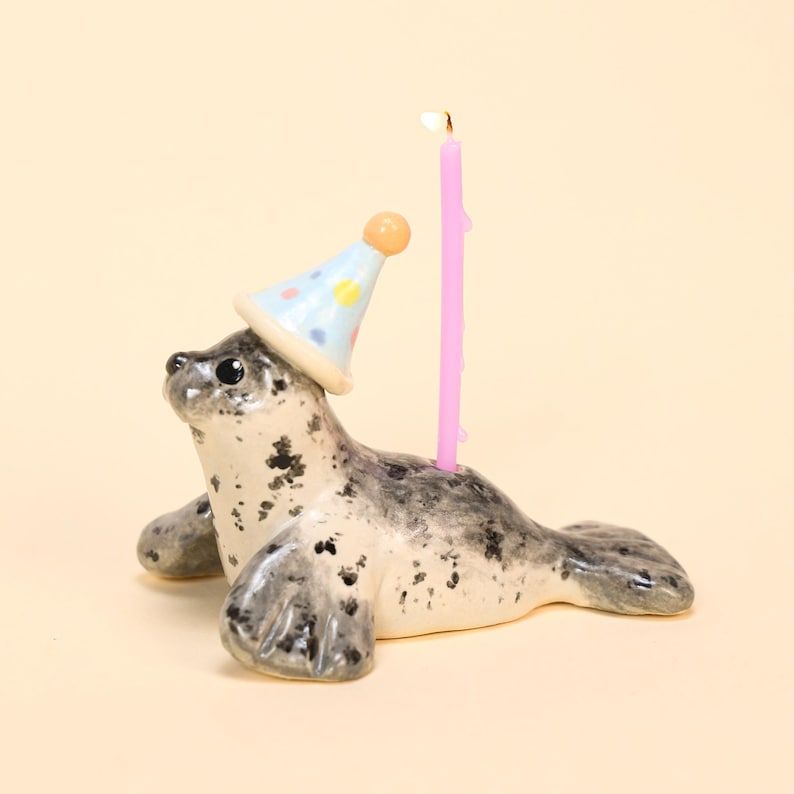 May include: A ceramic seal figurine with a light blue party hat adorned with colorful polka dots. A lit pink candle is placed on the seal's back. The seal has a speckled gray and white pattern.