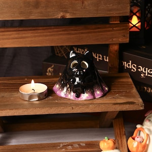 Pode incluir: Um castiçal preto em forma de gato de cerâmica com uma base roxa e dourada com padrão de estrelas. Uma vela de chá acesa está à esquerda do gato. O gato tem grandes buracos para os olhos e uma pequena figura de gato no interior.