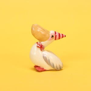 以下が含まれることがあります： 手描きのペリカンの置物。ペリカンは、大きな茶色のくちばし、ピンクと赤のストライプのくちばしの先端、赤い蝶ネクタイをしています。体はオフホワイトで灰色の翼のアクセントがあり、足は赤色です。背景は無地の黄色です。