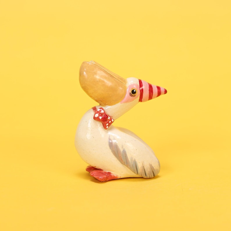 以下が含まれることがあります： 手描きのペリカンの置物。ペリカンは、大きな茶色のくちばし、ピンクと赤のストライプのくちばしの先端、赤い蝶ネクタイをしています。体はオフホワイトで灰色の翼のアクセントがあり、足は赤色です。背景は無地の黄色です。