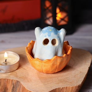 Peut inclure: Décoration d'Halloween en céramique. Un fantôme bleu clair avec une tête étoilée est assis dans une citrouille orange. La citrouille a une texture sculptée. Une bougie allumée est au premier plan, avec une lanterne sombre en arrière-plan.