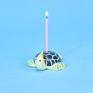 Puede incluir: Un original portavelas con forma de tortuga con una vela rosa encendida. La tortuga de cerámica tiene un esquema de color verde y blanco y un gorro de fiesta. El fondo es de color azul claro liso.