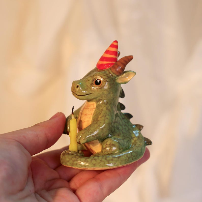 K&ouml;nnte beinhalten: Keramikfigur eines gr&uuml;nen Drachen mit roter und orangefarbener Partyhut. Der Drache h&auml;lt eine gelbe Kerze. Die Figur hat braune H&ouml;rner und eine detaillierte, strukturierte Oberfl&auml;che.