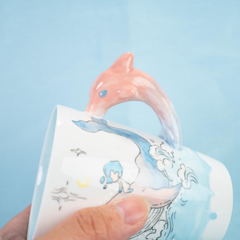 Puede incluir: Una taza de cer&aacute;mica blanca con un asa de delf&iacute;n rosa. La taza presenta una ilustraci&oacute;n acuarela azul y blanca de un delf&iacute;n saltando sobre una ola. La ilustraci&oacute;n tambi&eacute;n incluye una peque&ntilde;a persona en el agua.