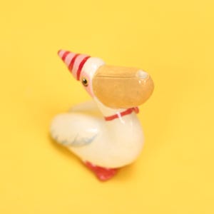 以下が含まれることがあります： パーティーハットをかぶったセラミック製のペリカンの置物。ペリカンは白で、茶色のくちばし、赤い足、赤い襟が付いています。パーティーハットは白地にピンクのストライプです。黄色い背景に配置されています。