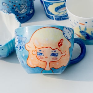May include: A blue ceramic mug with a hand-painted design of a girl with blonde hair and blue eyes. The girl is surrounded by blue and white fish and stars. The mug has a blue handle.